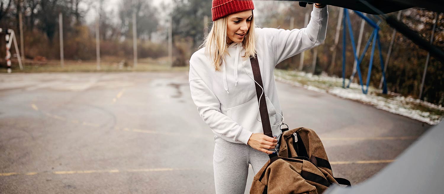 A blonde woman who knows how to wear sweatpants carrying a brown bag and closing the trunk of her car in a parking lot