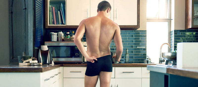 Man wearing only navy trunks facing a kitchen counter and grabbing a mug, with a couple fingers tucked into the back of his waistband