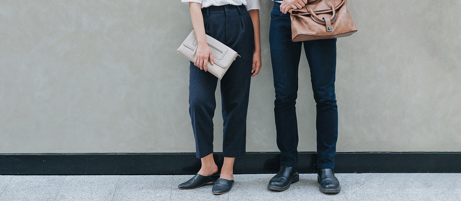 A woman wearing black ankle pants, black mule shoes, holding a gray clutch purse, and a man wearing tight black pants, black loafers, and holding a tan laptop bag are standing against a gray wall