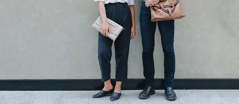 A woman wearing black ankle pants, black mule shoes, holding a gray clutch purse, and a man wearing tight black pants, black loafers, and holding a tan laptop bag are standing against a gray wall